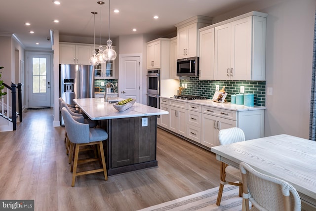 kitchen with stainless steel appliances, a center island with sink, tasteful backsplash, light wood-type flooring, and white cabinets