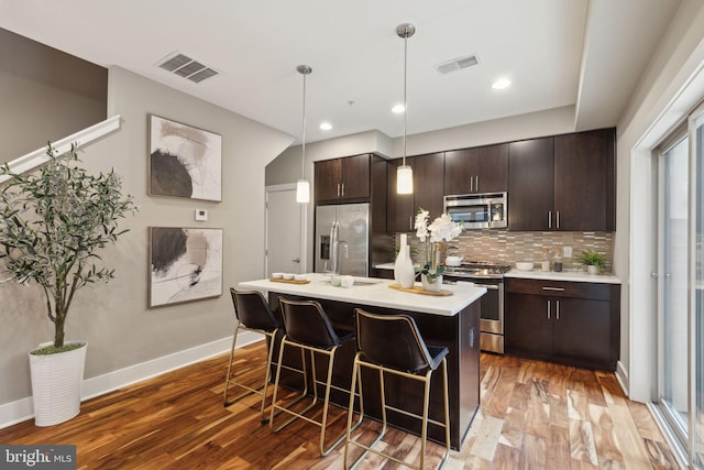 kitchen with dark brown cabinetry, hanging light fixtures, hardwood / wood-style floors, an island with sink, and appliances with stainless steel finishes