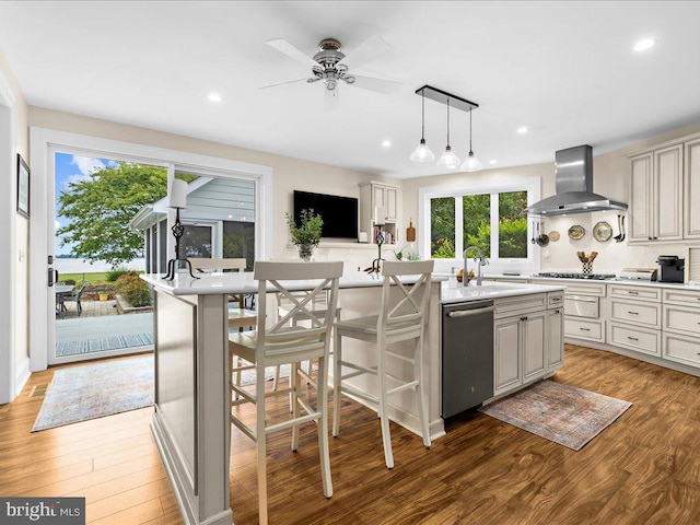 kitchen featuring hardwood / wood-style flooring, pendant lighting, wall chimney range hood, a kitchen island with sink, and appliances with stainless steel finishes