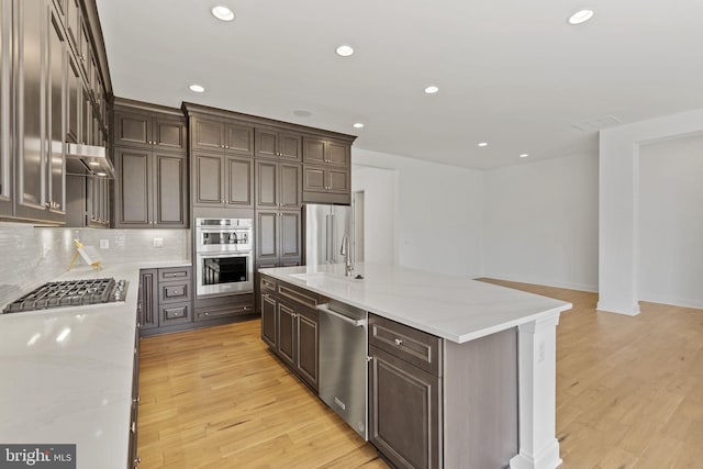 kitchen with light hardwood / wood-style floors, tasteful backsplash, an island with sink, light stone counters, and appliances with stainless steel finishes