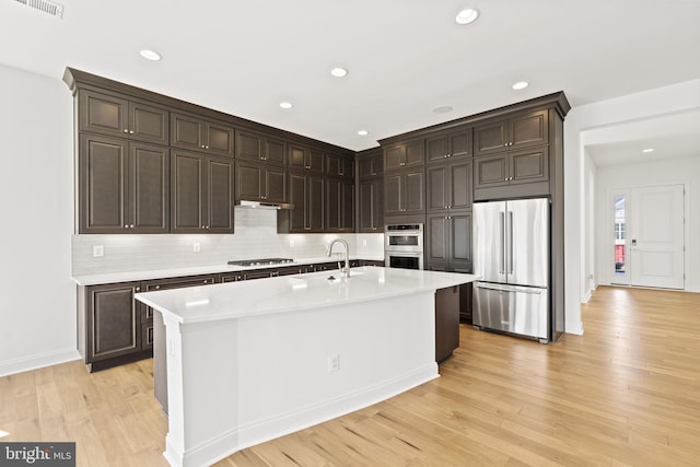 kitchen with appliances with stainless steel finishes, dark brown cabinetry, an island with sink, and light hardwood / wood-style flooring