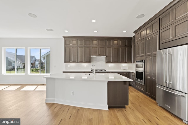 kitchen featuring light hardwood / wood-style floors, stainless steel appliances, a center island with sink, tasteful backsplash, and dark brown cabinets