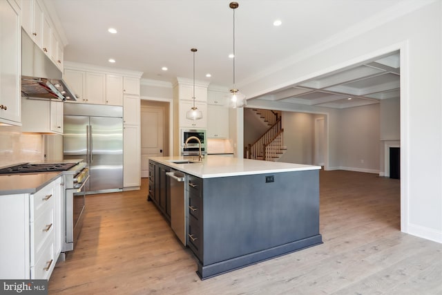 kitchen with stainless steel appliances, coffered ceiling, hanging light fixtures, and an island with sink