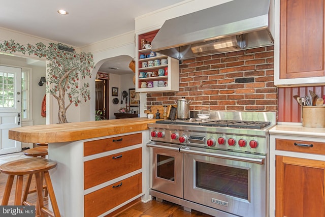 kitchen featuring wall chimney range hood, brick wall, range with two ovens, wood-type flooring, and a breakfast bar