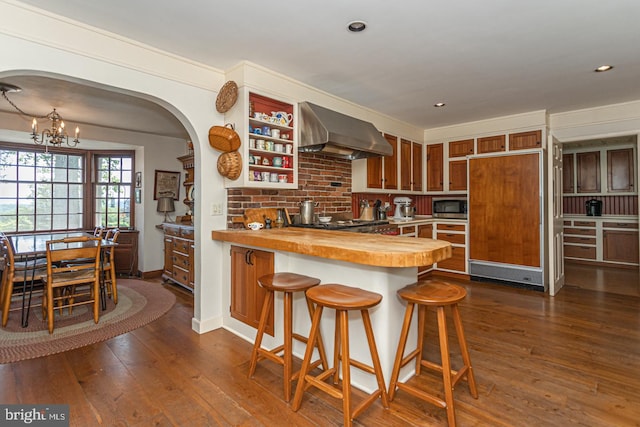 kitchen featuring wall chimney range hood, a notable chandelier, built in appliances, and dark wood-type flooring