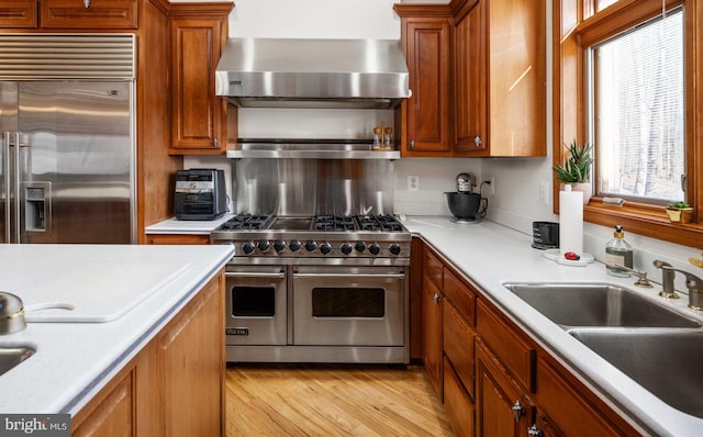 kitchen featuring wall chimney range hood, high quality appliances, light wood-type flooring, sink, and tasteful backsplash