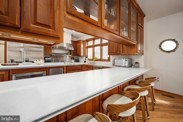 kitchen with light hardwood / wood-style flooring, kitchen peninsula, wall chimney range hood, a breakfast bar area, and appliances with stainless steel finishes