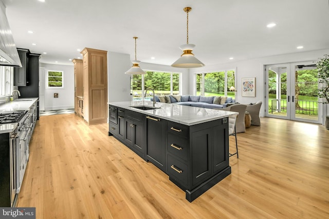 kitchen with light hardwood / wood-style floors, a kitchen island with sink, light stone countertops, sink, and pendant lighting