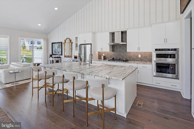 kitchen featuring dark hardwood / wood-style flooring, tasteful backsplash, a kitchen island with sink, wall chimney exhaust hood, and appliances with stainless steel finishes