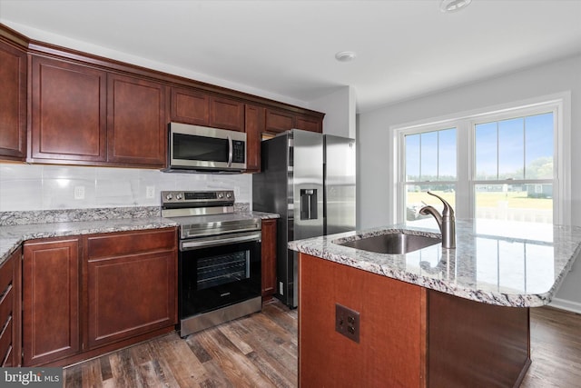 kitchen with dark wood-type flooring, a center island with sink, sink, light stone countertops, and stainless steel appliances