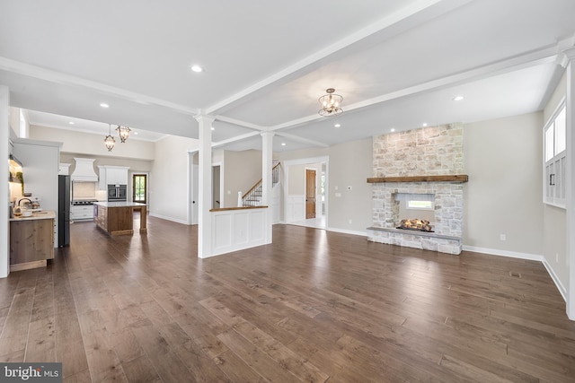 unfurnished living room featuring dark wood-type flooring, beamed ceiling, ornate columns, a stone fireplace, and a notable chandelier