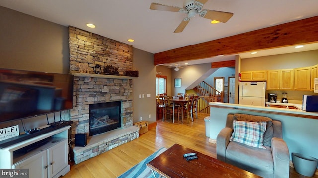 living room featuring beamed ceiling, light wood-type flooring, a stone fireplace, and ceiling fan