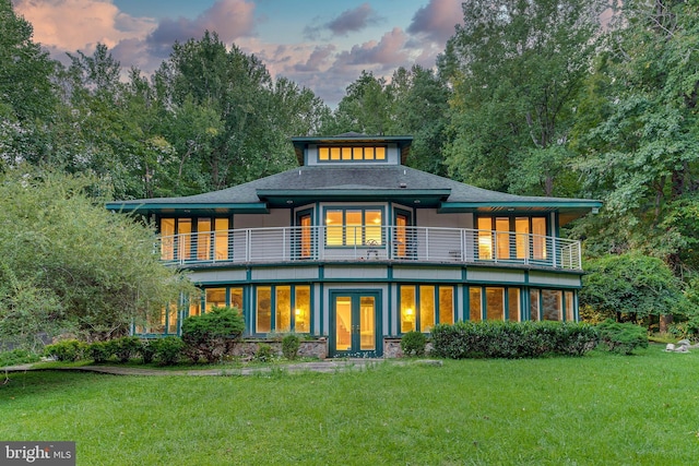 back of house at dusk featuring a balcony, french doors, and a yard