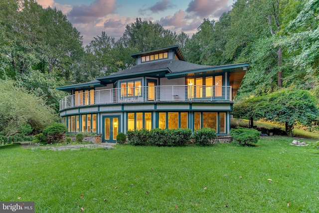 back of property at dusk featuring a balcony, a lawn, french doors, and a shingled roof