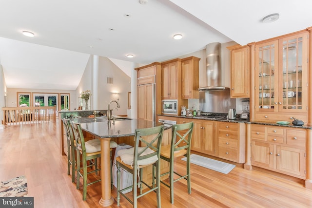 kitchen with stainless steel microwave, glass insert cabinets, wall chimney exhaust hood, and light brown cabinets