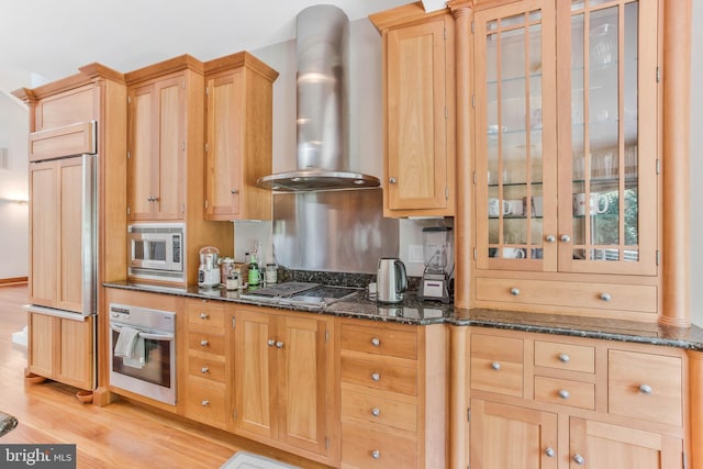 kitchen with light brown cabinets, built in appliances, dark stone countertops, light wood-style floors, and wall chimney exhaust hood