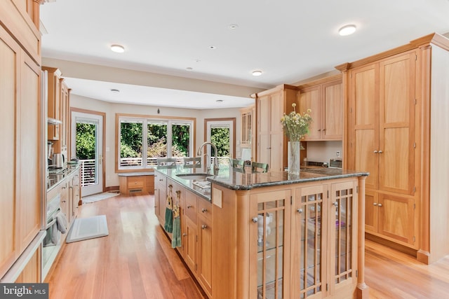 kitchen featuring a center island with sink, light wood finished floors, light brown cabinetry, a sink, and glass insert cabinets