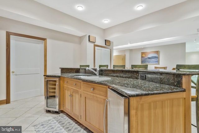 kitchen featuring light tile patterned floors, beverage cooler, dark stone counters, an island with sink, and a sink