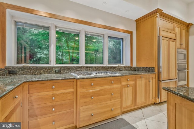 kitchen featuring dark stone countertops, built in appliances, brown cabinetry, and light tile patterned floors