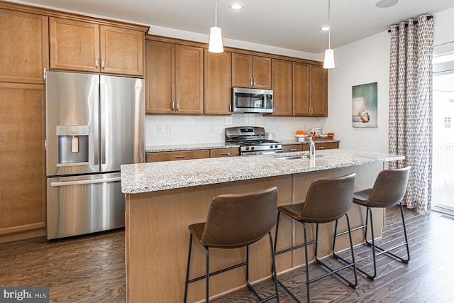 kitchen with hanging light fixtures, dark wood-type flooring, stainless steel appliances, a kitchen island with sink, and backsplash