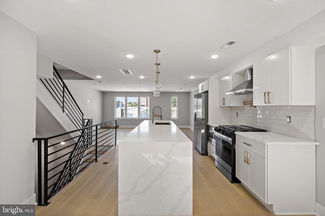 kitchen featuring appliances with stainless steel finishes, wall chimney exhaust hood, a kitchen island with sink, decorative light fixtures, and white cabinetry