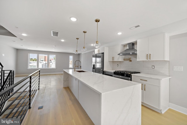 kitchen with white cabinetry, wall chimney exhaust hood, stainless steel appliances, light stone counters, and an island with sink