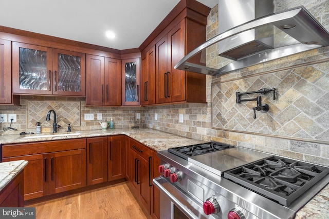 kitchen with wall chimney range hood, sink, stainless steel stove, light stone countertops, and light hardwood / wood-style floors