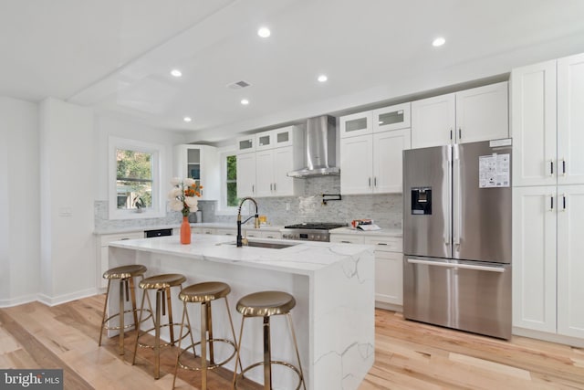 kitchen featuring wall chimney exhaust hood, light stone countertops, stainless steel appliances, and an island with sink