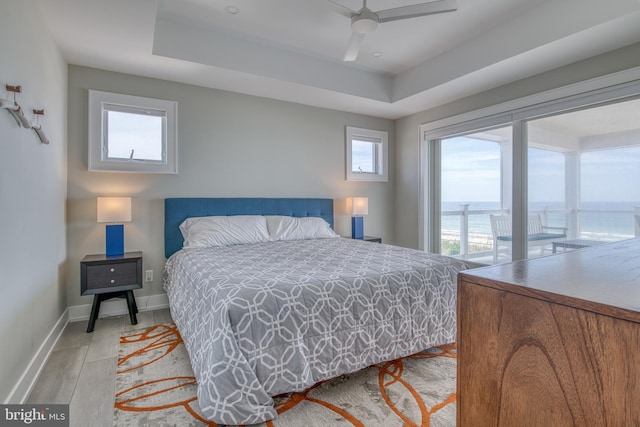 bedroom with access to exterior, ceiling fan, light hardwood / wood-style flooring, a tray ceiling, and a water view