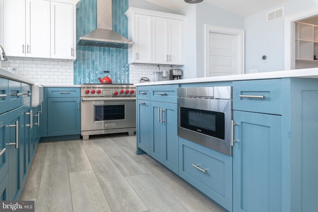 kitchen featuring white cabinetry, wall chimney range hood, light hardwood / wood-style flooring, decorative backsplash, and appliances with stainless steel finishes