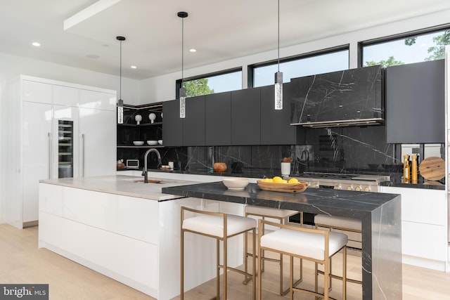 kitchen with hanging light fixtures, range hood, backsplash, light hardwood / wood-style floors, and a center island with sink