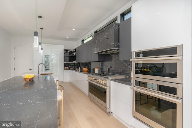 kitchen featuring tasteful backsplash, stainless steel appliances, wall chimney range hood, pendant lighting, and white cabinets