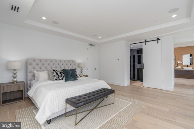 bedroom featuring a barn door, a tray ceiling, ensuite bath, and light hardwood / wood-style floors