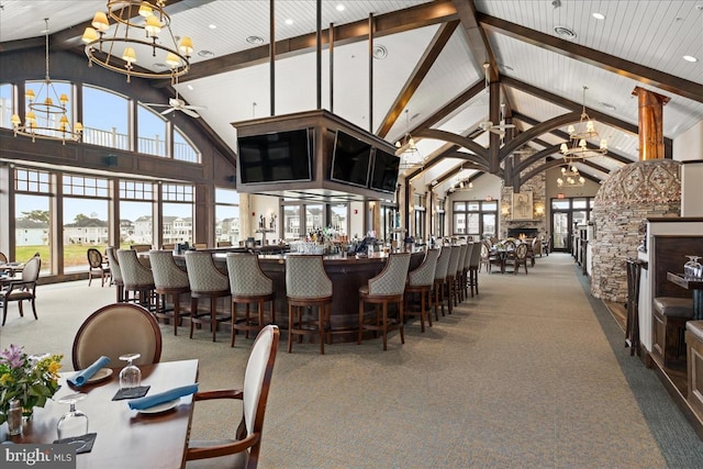 dining area featuring high vaulted ceiling, ceiling fan with notable chandelier, a stone fireplace, carpet flooring, and beam ceiling