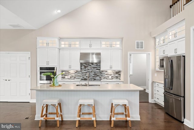 kitchen with wall chimney exhaust hood, stainless steel appliances, high vaulted ceiling, and an island with sink