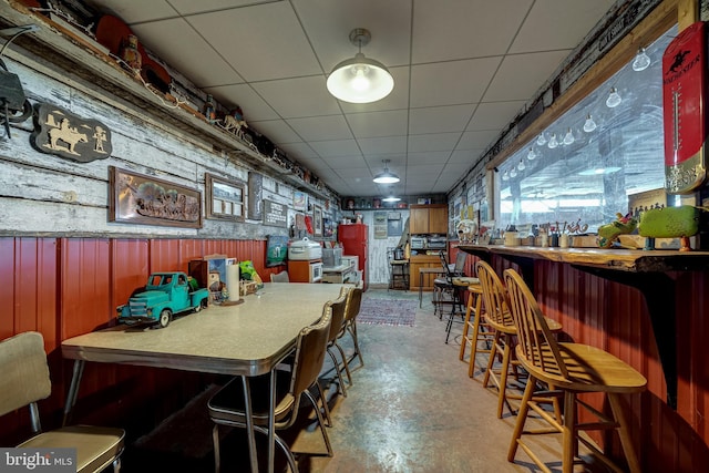 dining room featuring bar area, a paneled ceiling, wood walls, and concrete floors