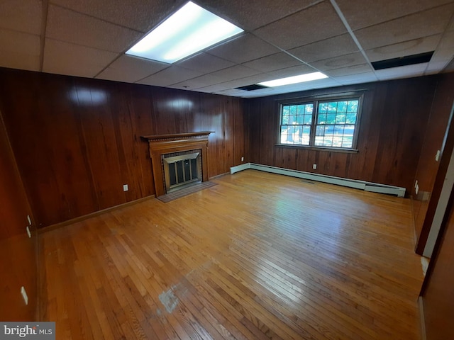 unfurnished living room with a paneled ceiling, wooden walls, light wood-type flooring, and a baseboard heating unit