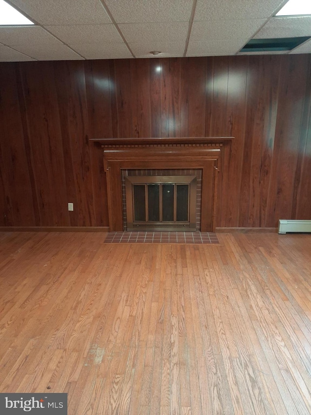 unfurnished living room with baseboard heating, wooden walls, a drop ceiling, and a brick fireplace