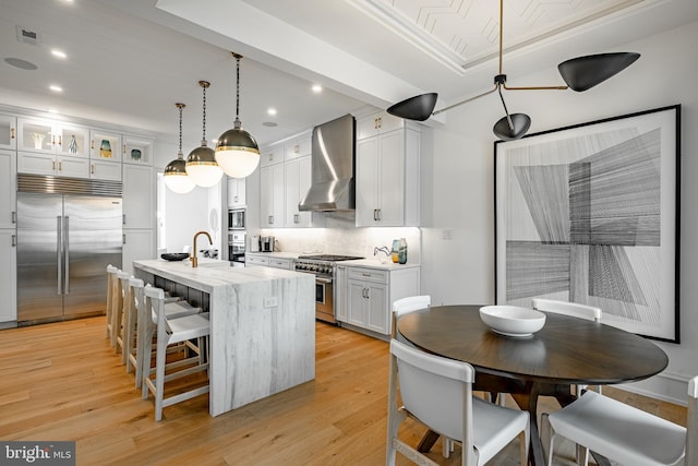 kitchen featuring white cabinetry, wall chimney range hood, built in appliances, light hardwood / wood-style floors, and a kitchen island with sink