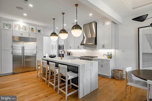 kitchen with light wood-type flooring, wall chimney range hood, pendant lighting, built in appliances, and a center island with sink