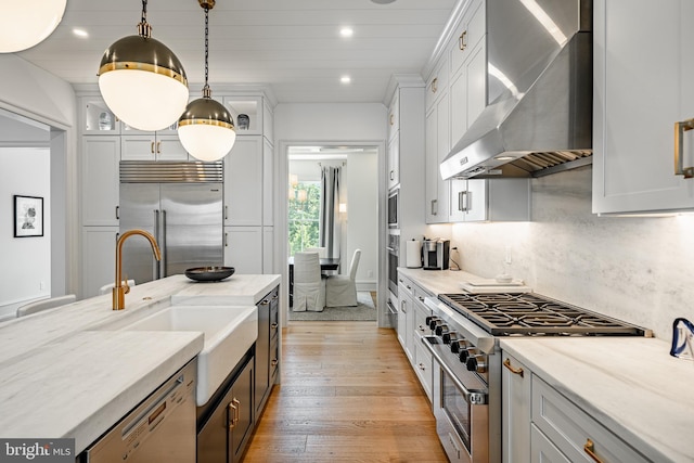 kitchen with high quality appliances, wall chimney range hood, light stone countertops, light wood-type flooring, and decorative light fixtures