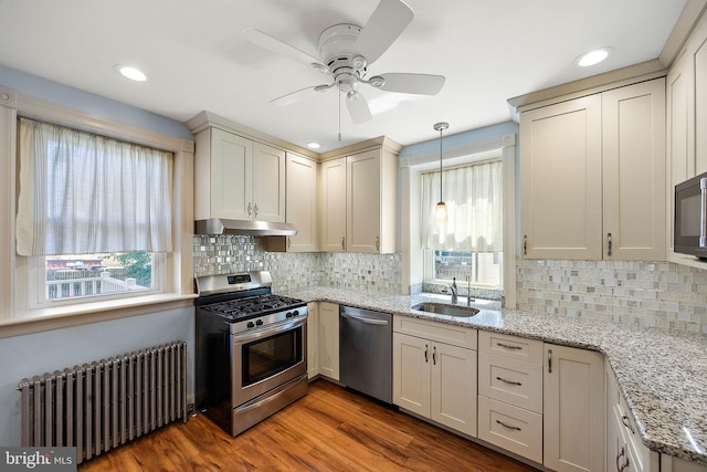 kitchen featuring appliances with stainless steel finishes, light stone counters, radiator, decorative light fixtures, and light hardwood / wood-style flooring