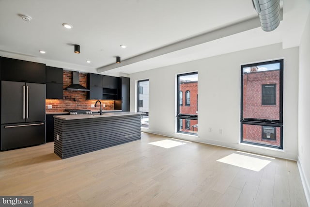 kitchen featuring backsplash, high end fridge, a kitchen island with sink, wall chimney range hood, and light hardwood / wood-style flooring