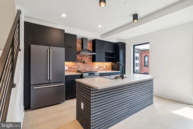 kitchen with sink, stainless steel appliances, wall chimney range hood, tasteful backsplash, and a center island with sink