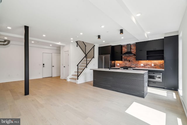 kitchen with appliances with stainless steel finishes, light wood-type flooring, backsplash, wall chimney exhaust hood, and a center island with sink