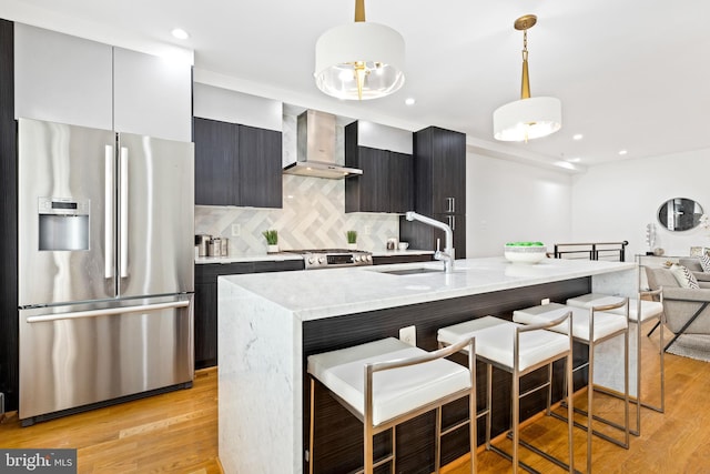 kitchen with appliances with stainless steel finishes, wall chimney exhaust hood, sink, decorative light fixtures, and light hardwood / wood-style floors