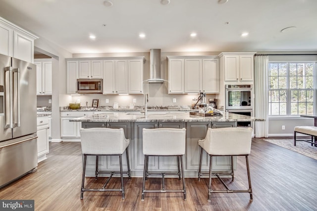 kitchen featuring a center island with sink, stainless steel appliances, wall chimney range hood, and light hardwood / wood-style flooring