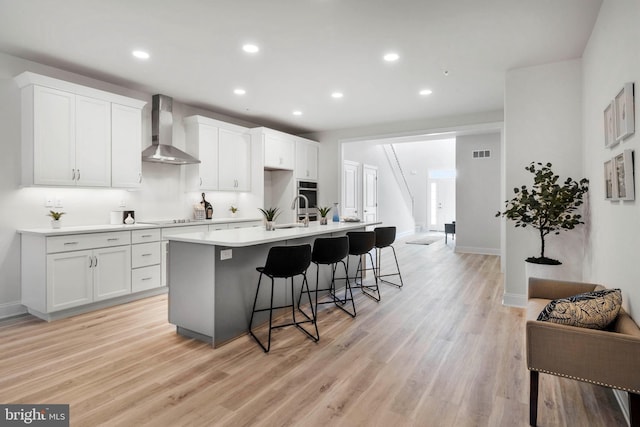 kitchen featuring a breakfast bar, a kitchen island with sink, white cabinets, wall chimney exhaust hood, and light wood-type flooring