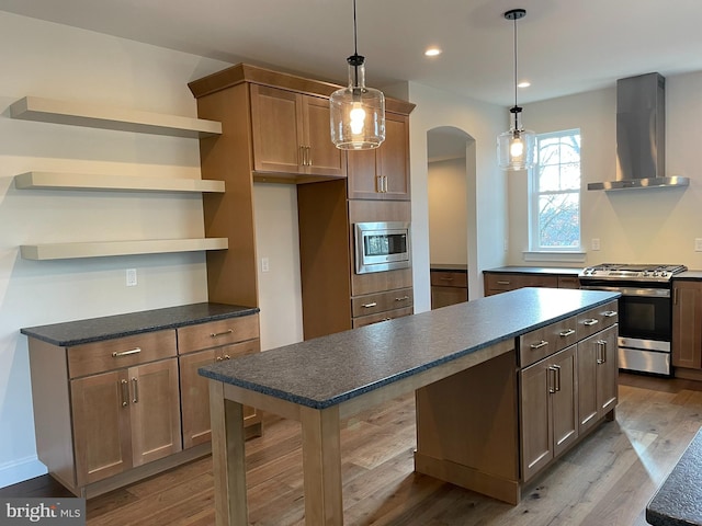 kitchen with pendant lighting, a center island, wall chimney range hood, light wood-type flooring, and stainless steel appliances