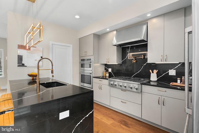 kitchen with sink, wall chimney range hood, decorative backsplash, appliances with stainless steel finishes, and light wood-type flooring